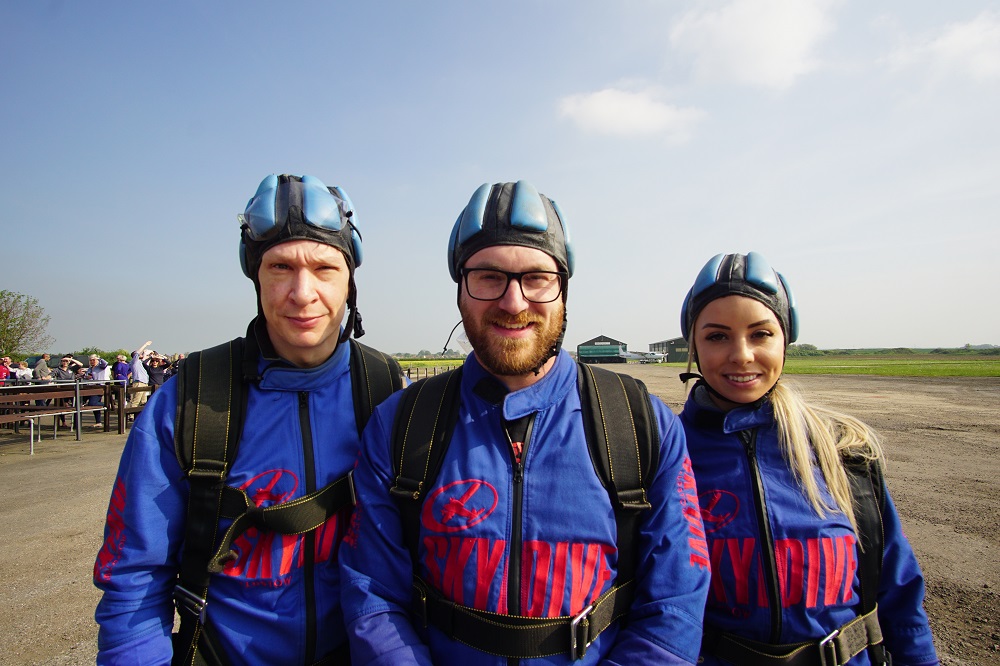 Three members of the Bytron team stand to pose for a photograph. They are wearing blue and red overalls and harnesses.  