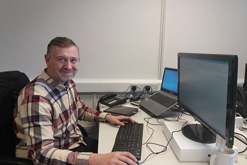 Pictured is Ross Aitkin, IT Support Manager, sat at his desk smiling to the camera. On his desk is a laptop and separate screen, keyboard and mouse.